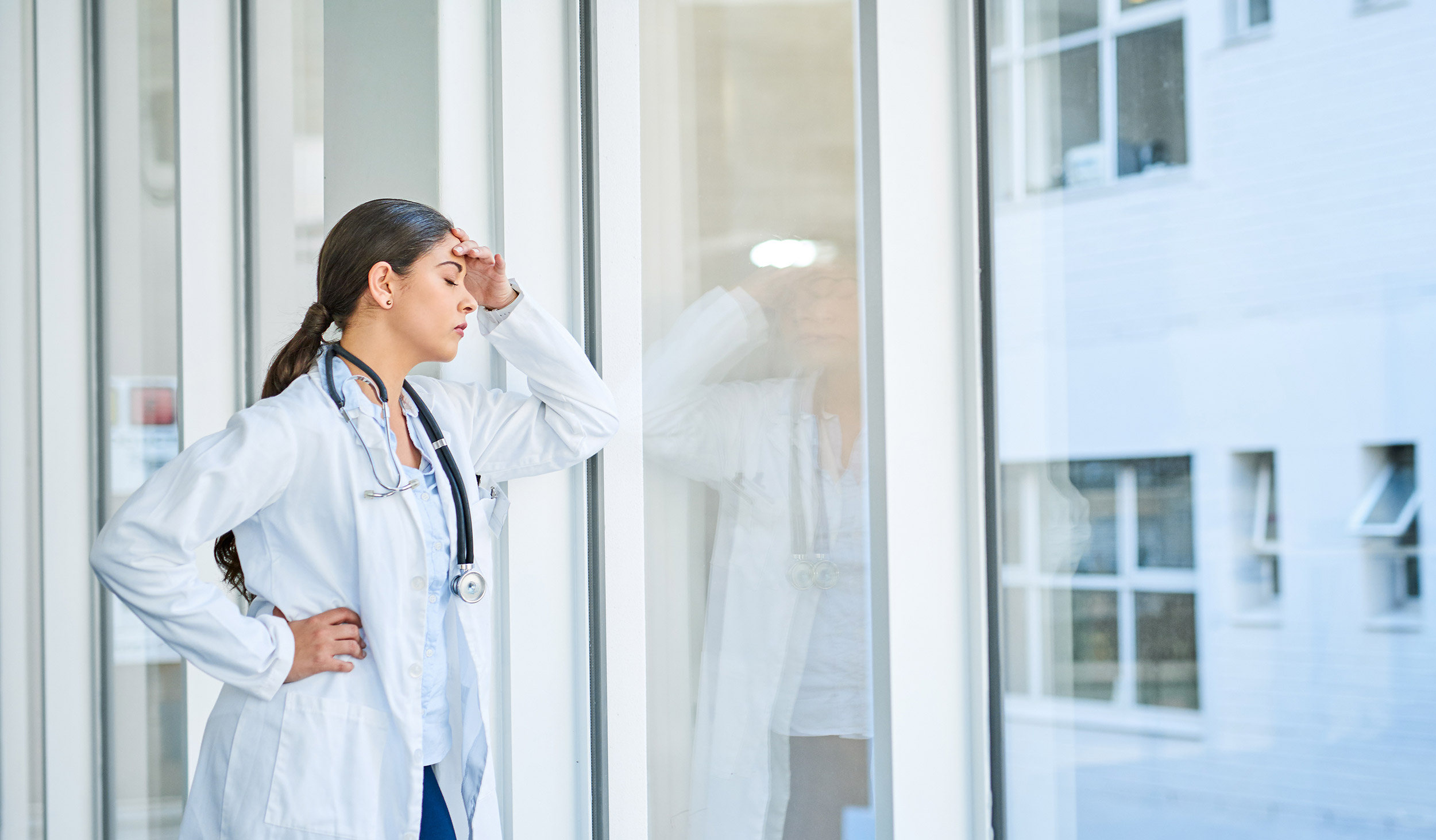A white female physician whose eyes are closed with her hand over her forehead  and appears to be stressed facing a large window. 